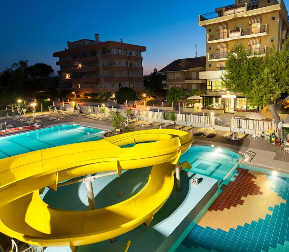 Vista serale della piscina con acquascivolo dell’Hotel Caravel B&B, illuminata e circondata da lettini.